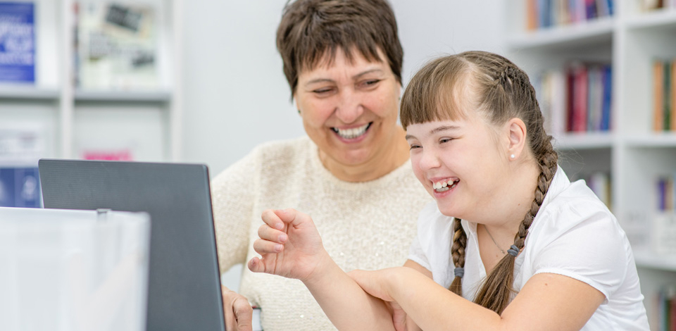 Disability worker smiling at young girl with Down Syndrome using laptop.
